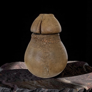 Op de afbeelding: Een gesneden houten vat met een bolvormig lichaam en een afgerond deksel. Het vat is lichtbruin van kleur met zichtbare houtnerf en een getextureerde band nabij de bovenkant. De achtergrond is zwart en het vat staat op een donker, getextureerd oppervlak.