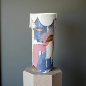 Könnte beinhalten: Eine hohe, zylindrische Keramikvase mit weißem, gewelltem Rand. Die Vase ist mit abstrakten Strichen in Rosa, Blau und Gold bemalt. Die Vase steht auf einem hellgrauen, sechseckigen Sockel.