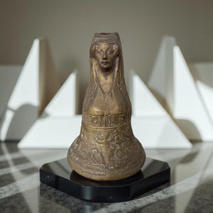 May include: A bronze-colored sculpture of a woman wearing a hooded garment. The sculpture has intricate details, including facial features and decorative patterns. It sits on a black square base, with a blurred white geometric background.