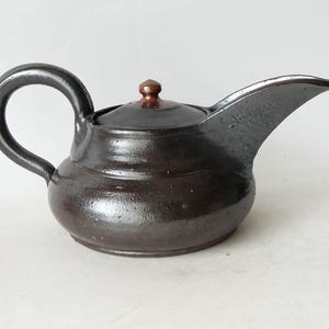 May include: A dark brown ceramic teapot with a rounded body, a curved handle, and a spout. The lid is a darker brown color. The teapot has a rustic, handmade appearance, suggesting it is a piece of pottery.