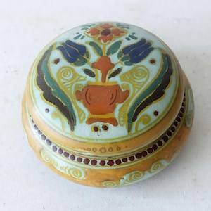 May include: A small, round, ceramic trinket box with a lid. The lid is decorated with a floral design in shades of blue, green, yellow, and orange. The box is decorated with a yellow and brown pattern.