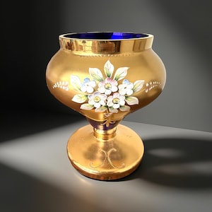 Op de afbeelding: Een goudkleurige decoratieve vaas met een blauw interieur. De vaas heeft handgeschilderde witte en roze bloemen met groene bladeren. De basis heeft een cirkelvormig ontwerp met gouden accenten. De vaas is een decoratief item.