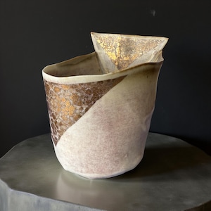 以下が含まれることがあります： ユニークな非対称デザインの手作りセラミック花瓶。花瓶は、片側にテクスチャ加工された茶色と金のパターン、もう片側に柔らかいオフホワイトの仕上げが施された対角カラーブロックが特徴です。上部には折り畳まれた三角形のアクセントがあります。