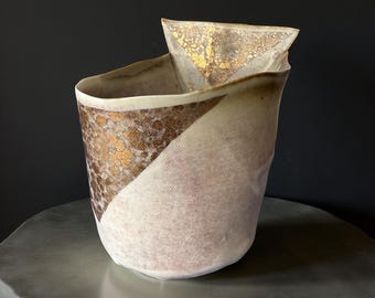 May include: A handcrafted ceramic vase with a unique, asymmetrical design. The vase features a diagonal color block, with a textured brown and gold pattern on one side and a soft, off-white finish on the other. The top has a folded, triangular accent.