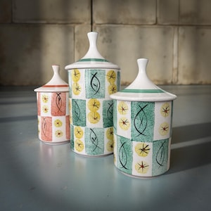 Op de afbeelding: Drie keramische potten met deksels. De potten zijn cilindrisch met een witte basis en deksel, en een groene streep rond het deksel. De potten hebben een geruit patroon met gele en zwarte ontwerpen. Eén pot is rood.