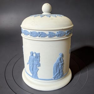 Op de afbeelding: Een cilindrische witte stenen pot met deksel, voorzien van lichtblauwe reliëfdecoraties. Het ontwerp bevat een rand van laurierbladeren aan de bovenkant en klassieke figuren, waaronder een persoon met een lier, op de romp.