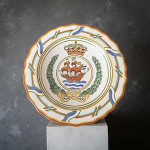 Puede incluir: Plato de cerámica decorativo con borde festoneado. El plato presenta un emblema central de un barco, corona y corona de laurel, con las fechas 1856 y 1936. El borde está decorado con detalles en azul, verde y naranja.