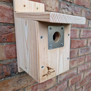 May include: A wooden birdhouse with a metal entrance hole and a sloping roof. The birdhouse is mounted on a brick wall and has the text "Longton Nature Reserve" printed on the front.