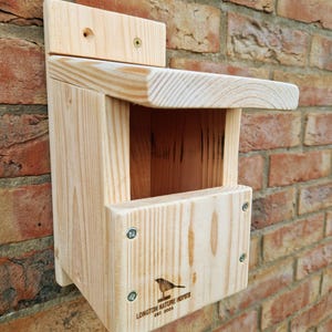 May include: A wooden birdhouse with a slanted roof and a small opening for birds to enter. The birdhouse is mounted on a brick wall and has a small sign on the bottom that reads "Longton Nature Homes Est. 2004".