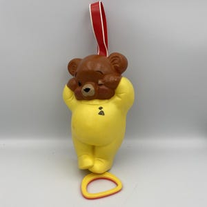 May include: A vintage yellow bear-shaped ornament with a brown head and a red ribbon for hanging. The bear is wearing a yellow outfit and has its hands up near its face. A yellow and red ring is at the bottom.