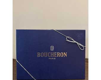 May include: A rectangular dark blue gift box featuring the gold embossed text BOUCHERON PARIS and a small logo above it. A thin white string is tied around the box in a decorative manner, forming a small bow on the upper right side of the lid.