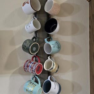 May include: A wooden mug rack with hooks holds a variety of mugs with different designs and colors. The mugs include a green mug with a tree design, a blue mug with the text "We also love our others", a white mug with the text "Snuggle", a red and white mug with a geometric pattern, a blue mug with the text "It's the little things that make you a queen", a black mug, a white mug with a floral design, and a white mug with a brown and white dog design.