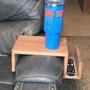 Op de afbeelding: Een houten armleuningblad met een plat oppervlak en een zijvak met een zwarte afstandsbediening. Een blauwe roestvrijstalen beker met een bruin rietje en een oranje grafisch logo met een gevleugeld wezen en de tekst ST staat op het blad.