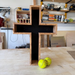 May include: A large wooden cross with a dark interior and a light brown exterior. Two yellow softballs are resting at the base of the cross. The cross is displayed on a light-colored wooden surface.