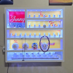 Op de afbeelding: Een wit wandrek met geïntegreerde ledverlichting, voorzien van meerdere horizontale niveaus met kleine pinnetjes voor het opbergen van honkballen. De unit bevat een rood paneel met de naam Jenny en gele achterwanden met honkbalafbeeldingen.
