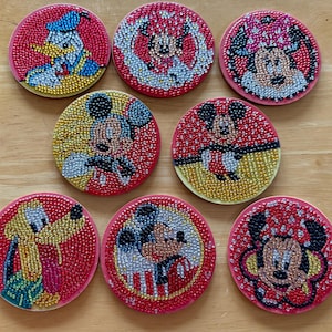 May include: Set of eight round coasters featuring Disney characters like Mickey Mouse, Minnie Mouse, Donald Duck, and Pluto. Each coaster has a red background with a beaded design, and a light-colored border.