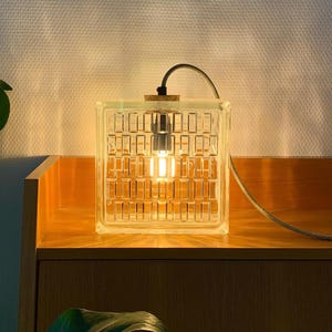 May include: A clear glass block lamp with a grid pattern, illuminated by a warm light bulb. The lamp sits on a wooden surface, with a gray cord extending from the top. The design is modern and minimalist.