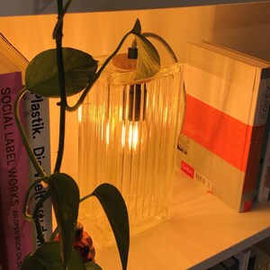 Może przedstawiać: Przezroczysta lampa szklana z ciepłą żar&oacute;wką, stojąca na p&oacute;łce z książkami. Lampa ma fakturowany, żebrowany wz&oacute;r i korkową podstawę.