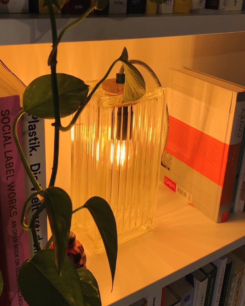 Może przedstawiać: Przezroczysta lampa szklana z ciepłą żar&oacute;wką, stojąca na p&oacute;łce z książkami. Lampa ma fakturowany, żebrowany wz&oacute;r i korkową podstawę.