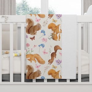 May include: A baby blanket with a repeating pattern of watercolour squirrels, butterflies, and flowers in brown, pink, blue, and purple on a cream background. The blanket is displayed on a white cot.