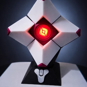 Puede incluir: Una escultura geométrica blanca y roja con un centro rojo brillante y una base negra. La escultura tiene forma de diamante con detalles rojos y un emblema plateado en la base. El objeto está sobre una superficie blanca.