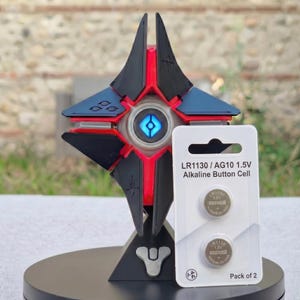 May include: A black, red, and blue geometric sculpture with a glowing blue center, resembling a futuristic design. Next to it is a package of two LR1130/AG10 1.5V alkaline button cell batteries. The package has the text "Pack of 2".