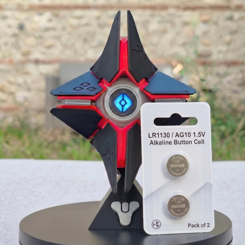 May include: A black, red, and blue geometric sculpture with a glowing blue center, resembling a futuristic design. Next to it is a package of two LR1130/AG10 1.5V alkaline button cell batteries. The package has the text "Pack of 2".