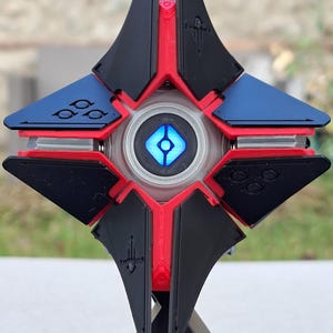 May include: A detailed, multi-colored 3D-printed Ghost from the video game Destiny. The Ghost features a black, red, and blue color scheme with a glowing blue center. It is mounted on a black stand with a white symbol.