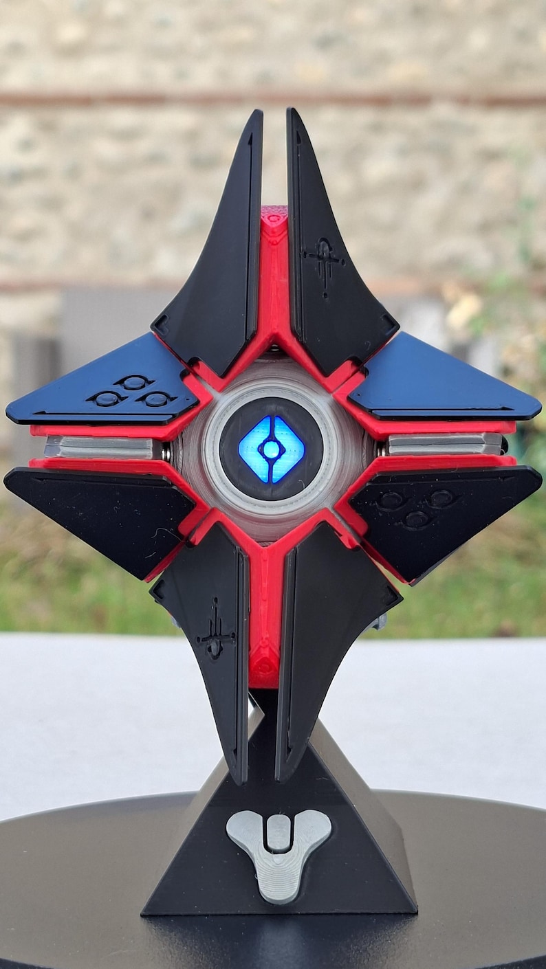 May include: A detailed, multi-colored 3D-printed Ghost from the video game Destiny. The Ghost features a black, red, and blue color scheme with a glowing blue center. It is mounted on a black stand with a white symbol.