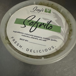 May include: A round, clear plastic container of Jay's Latin Kitchen sofrito. The label features the word "Sofrito" in cursive, with a green stripe and the ingredients listed below. The words "Fresh. Delicious." are also printed on the label.