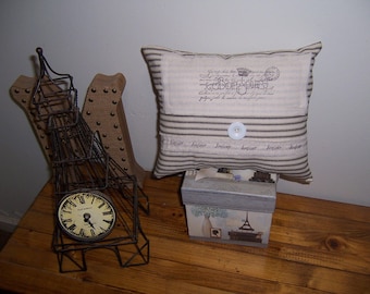 May include: A decorative pillow with a white background and brown stripes. The pillow has a vintage look and features the text "Bonjour" in a script font. The pillow is sitting on a wooden table with a metal Eiffel Tower replica and a clock.