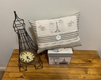 May include: A decorative scene featuring a metal Eiffel Tower clock, a striped pillow with Eiffel Tower imagery, and a small box. The pillow has a button closure and the clock has a cream-colored face. The items are on a wooden surface.