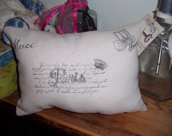 May include: A beige pillow with a black and white print of a Parisian scene. The pillow has the word "Merci" printed on it in black. The pillow is sitting on a wooden surface.