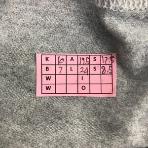 Pu&ograve; includere: Primo piano di un tessuto grigio con un'etichetta rettangolare rosa. L'etichetta presenta una griglia di caselle con numeri e lettere, tra cui "10", "19.5", "17.5", "7" e "24".