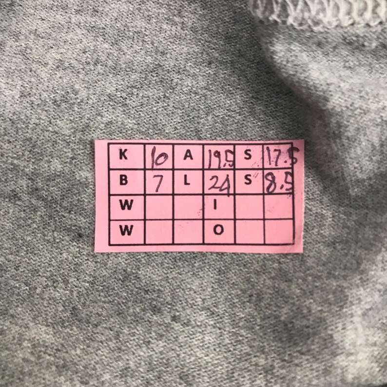 Pu&ograve; includere: Primo piano di un tessuto grigio con un'etichetta rettangolare rosa. L'etichetta presenta una griglia di caselle con numeri e lettere, tra cui "10", "19.5", "17.5", "7" e "24".