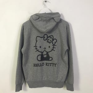 May include: Grey zip-up hoodie with a black Hello Kitty graphic wearing a skull bow, and the text "HELLO KITTY". The hood has ear-shaped details. A casual, comfortable garment.