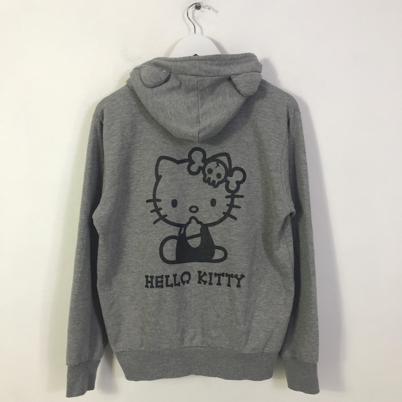 May include: Grey zip-up hoodie with a black Hello Kitty graphic wearing a skull bow, and the text "HELLO KITTY". The hood has ear-shaped details. A casual, comfortable garment.