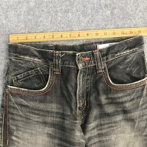May include: Close-up of a pair of faded black denim jeans. The jeans have orange stitching along the pockets and seams. The jeans have a button closure and a small coin pocket. The jeans are laid flat on a gray surface.