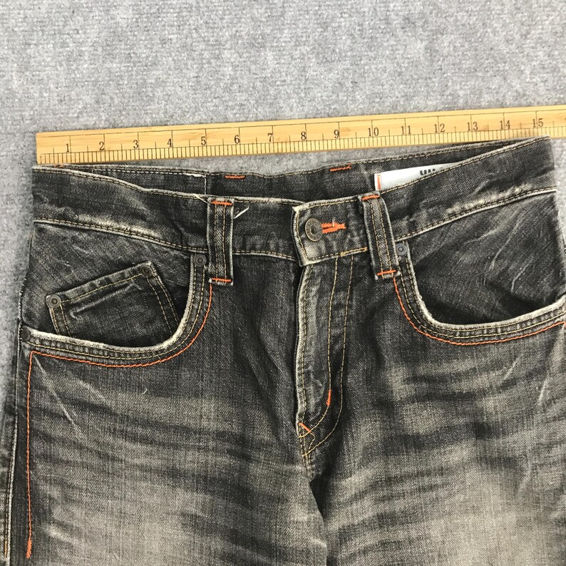 May include: Close-up of a pair of faded black denim jeans. The jeans have orange stitching along the pockets and seams. The jeans have a button closure and a small coin pocket. The jeans are laid flat on a gray surface.