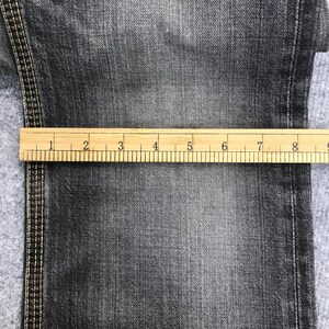 May include: Close-up of dark gray denim fabric with a wooden ruler measuring up to 9 inches. The denim has visible texture and stitching along the edge. The ruler is marked with inch increments.