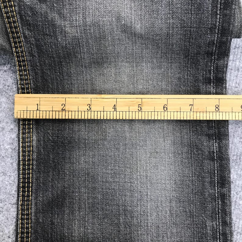 May include: Close-up of dark gray denim fabric with a wooden ruler measuring up to 9 inches. The denim has visible texture and stitching along the edge. The ruler is marked with inch increments.