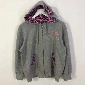 May include: A gray zip-up hoodie featuring a Hello Kitty design with a pink bow and gold outline. The hood lining and pocket accents have a pink and black leopard print. The hood has cat ear details.