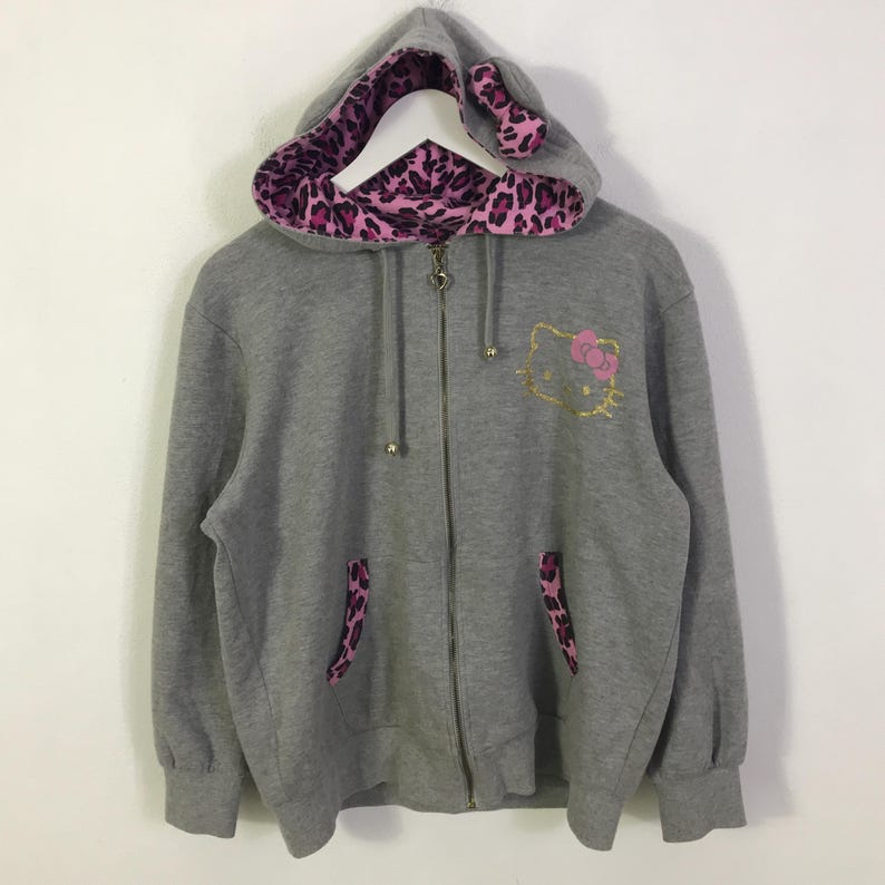 May include: A gray zip-up hoodie featuring a Hello Kitty design with a pink bow and gold outline. The hood lining and pocket accents have a pink and black leopard print. The hood has cat ear details.