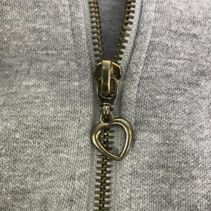 May include: Close-up of a gray hoodie zipper with a gold-colored metal zipper pull in the shape of a heart. The zipper is partially closed, revealing the teeth and the fabric of the hoodie.