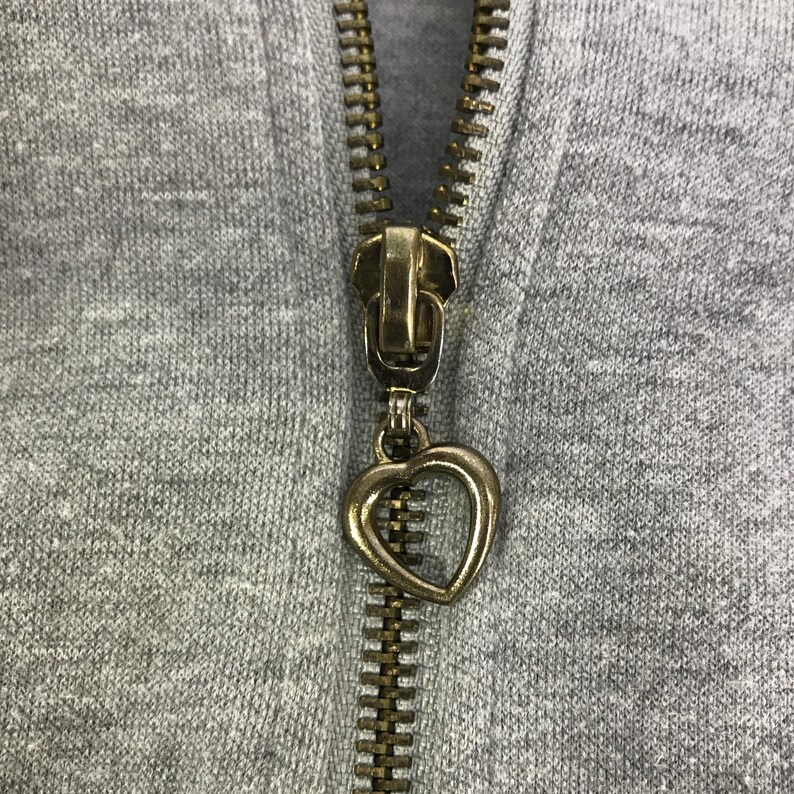 May include: Close-up of a gray hoodie zipper with a gold-colored metal zipper pull in the shape of a heart. The zipper is partially closed, revealing the teeth and the fabric of the hoodie.