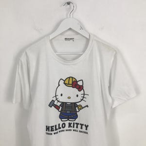 May include: White t-shirt featuring a cartoon image of Hello Kitty dressed as a construction worker. The text "Hello Kitty Those who work hard will succeed." is printed below the image.