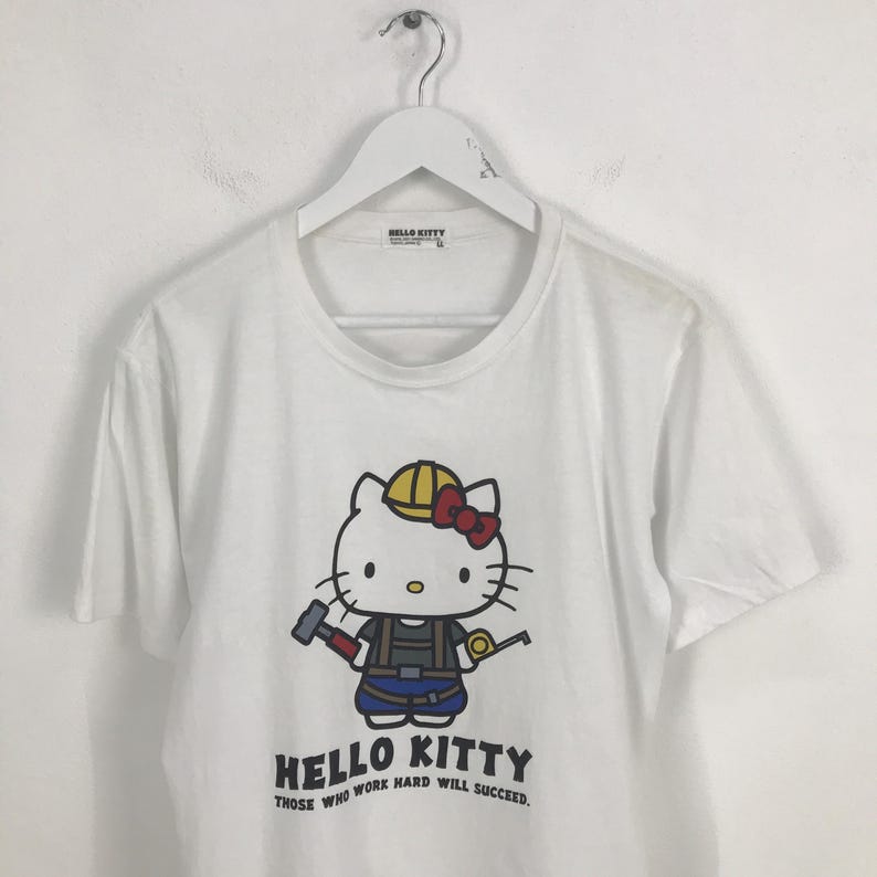May include: White t-shirt featuring a cartoon image of Hello Kitty dressed as a construction worker. The text "Hello Kitty Those who work hard will succeed." is printed below the image.