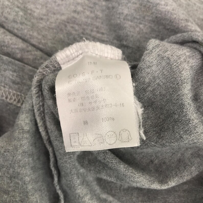 Pu&ograve; includere: Primo piano di un capo di abbigliamento in tessuto grigio con un'etichetta bianca. L'etichetta contiene testo tra cui "CO/S-P-T" e "GANRIO &copy;". Il tessuto &egrave; piegato e l'etichetta &egrave; attaccata al colletto del capo. Sono visibili anche le istruzioni di lavaggio.