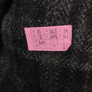 May include: Close-up of dark grey fabric with a pink tag attached. The tag has handwritten numbers and letters in black ink. The fabric appears to be a textured knit or woven material.