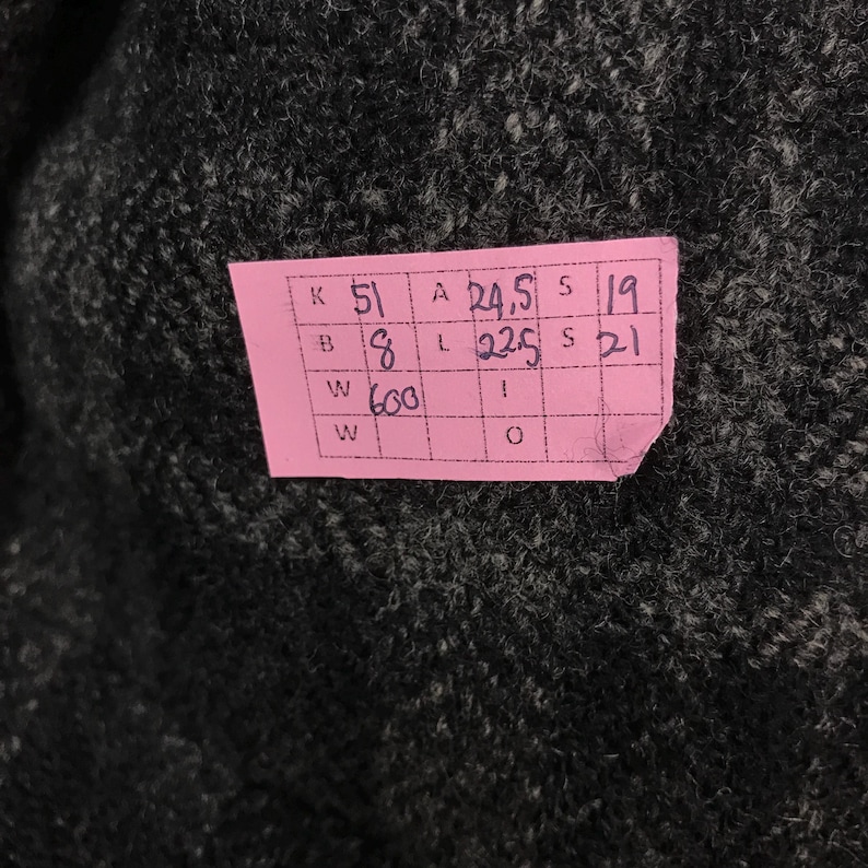 May include: Close-up of dark grey fabric with a pink tag attached. The tag has handwritten numbers and letters in black ink. The fabric appears to be a textured knit or woven material.